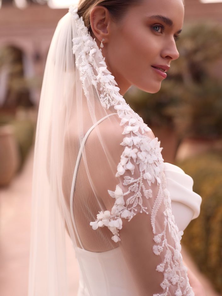 Eine Braut in einem weißen Kleid und einem Schleier aus Blumenspitze steht im Freien, blickt über die Schulter und präsentiert ihr elegantes Brautoutfit.