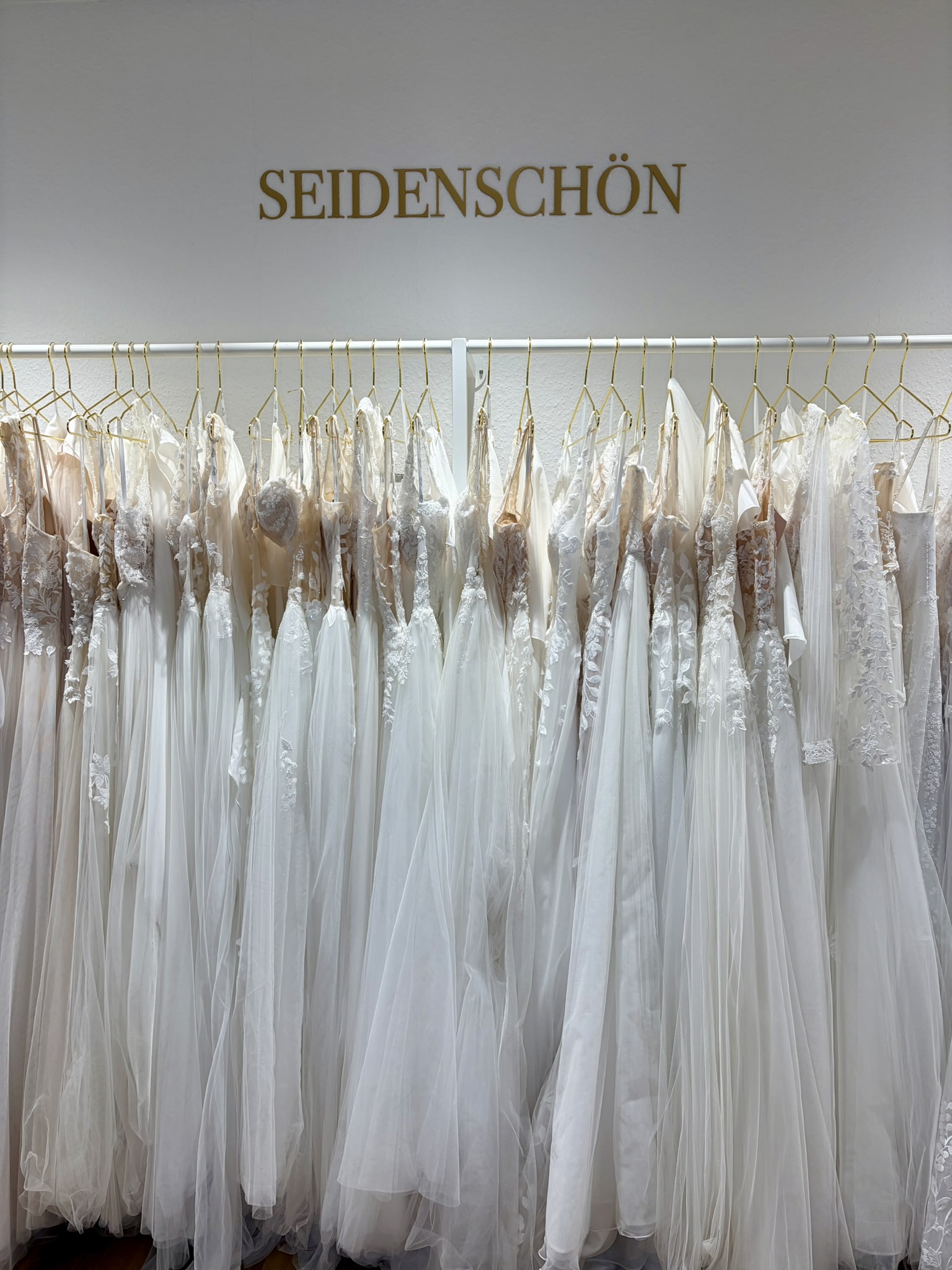Eine Reihe weißer Brautkleider auf goldenen Kleiderbügeln unter einem Schild mit der Aufschrift „SEIDENSCHÖN“ – jedes einzelne wartet darauf, von seiner perfekten Braut entdeckt zu werden, die in diesem eleganten Brautmodengeschäft ihr Traumoutfit findet.
