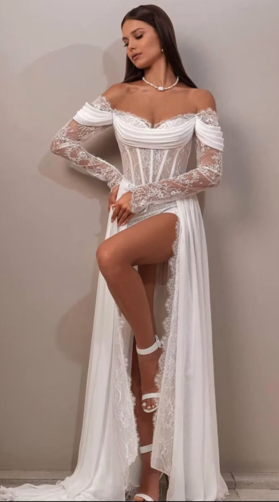 Eine Braut in einem schulterfreien weißen Spitzenkleid mit hohem Schlitz, dazu weiße Pumps und eine Perlenkette – ein elegantes Brautoutfit, das perfekt in jeden Brautmodenladen passt.