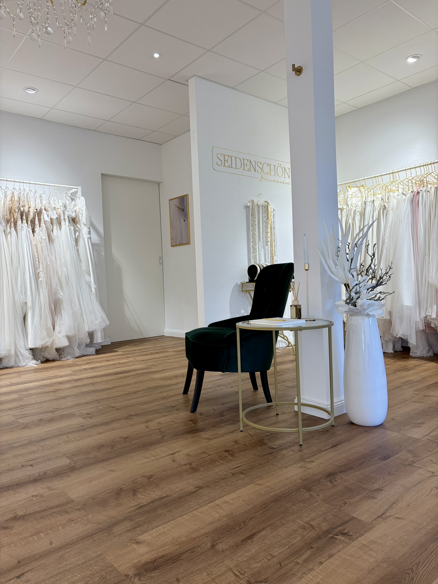 Eine Brautmodenboutique mit eleganten Brautkleidern an Kleiderständern, einem grünen Stuhl, einem kleinen Tisch und einer weißen Vase auf einem Holzboden.