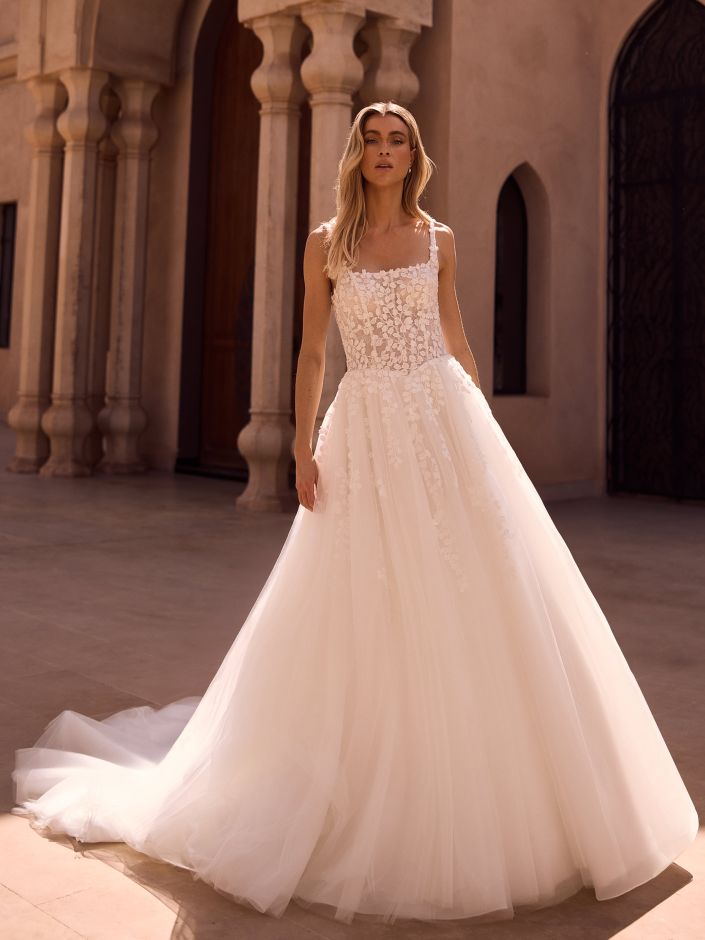 Eine Frau in einem weißen Brautkleid aus Blumenspitze steht vor einem Gebäude mit gewölbten Türen.