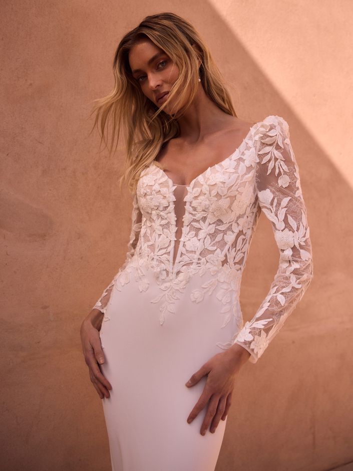 Eine Frau posiert in einem langärmeligen Brautkleid aus Spitze mit floralen Applikationen vor einer pfirsichfarbenen Wand und präsentiert damit ein elegantes Brautoutfit, das perfekt für jede moderne Braut ist.
