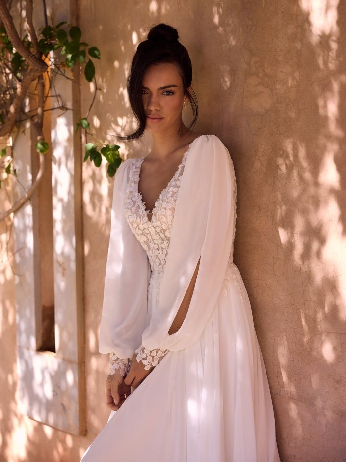 Eine Frau in einem langärmeligen, weißen Spitzen-Brautkleid steht vor einer sonnendurchfluteten, strukturierten Wand mit Grünpflanzen im Hintergrund und verkörpert die Eleganz eines perfekten Brautoutfits.